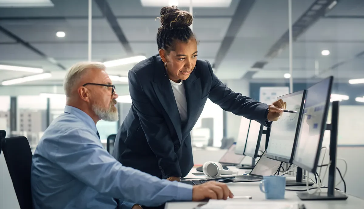 Compliance by Design: Building Resilience That Lasts // Black female manager pointing to desktop computer screen operated by male worker in modern office